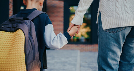 Posterior camera view of little Caucasian boy with blond hair walking towards school together with...