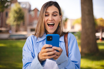 Blonde short hair woman browsing internet with cellphone mobile phone wearing casual clothes shirt gesturing like winner. Smiling open mouth woman look happy, celebrating win, triumph, success, wow.
