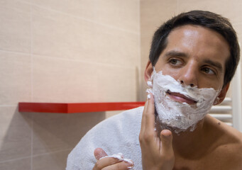 Close-up of a handsome young man applying shaving foam to his face while looking in the bathroom...