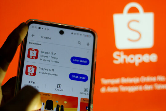 pringsewu, Lampung; November 27, 2024; man's hand holding a selective cell phone screen with two shopee 12.12 application options blurring the computer monitor on the official Shopee Indonesia website