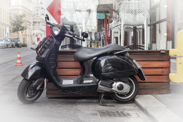 A black scooter is parked on a city street near an outdoor cafe