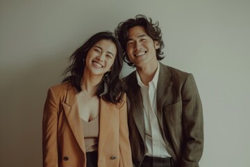 Portrait of a cheerful asian couple in their 20s dressed in a stylish blazer while standing against bare monochromatic room