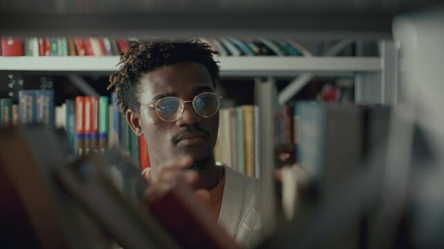 Young Black male student in glasses looking for book on shelf in college library, examining covers and checking text