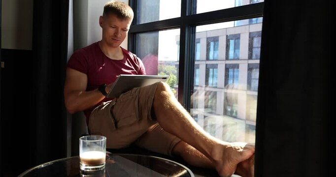 Guy freelancer sits on windowsill and works on project using tablet. Young man uses contemporary tablet to study graphs and edit documents