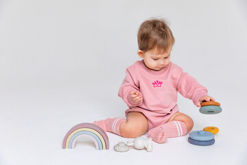 Happy smiling little baby girl sits on the floor and plays with a multi-colored pyramid sorter and rainbow toy on a white background. Development of fine motor skills. Early education. Copyspace