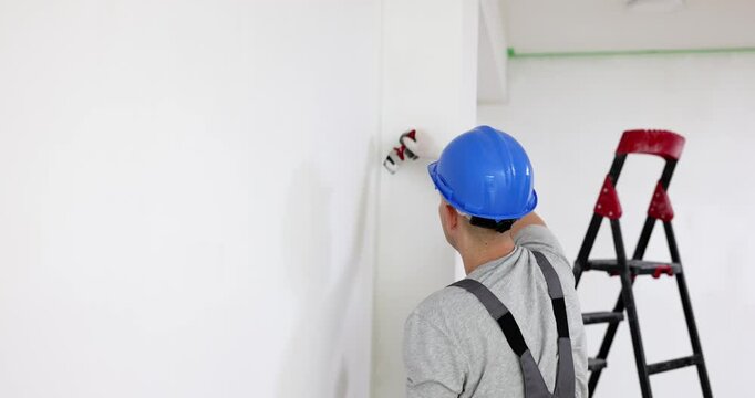 Man builder in helmet paints wall with brush using white color against ladder. Careful construction worker in uniform engages in renovation process