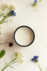 Black jar with cream surrounded by flowers on wooden table, top view