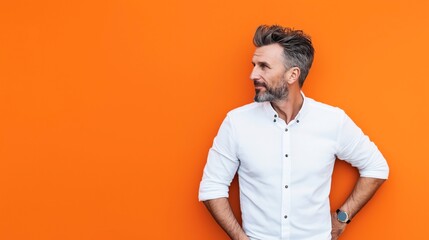 Fototapeta premium Middle-aged man with gray beard and stylish hair wearing a white shirt, standing against an orange background, looking to the side.
