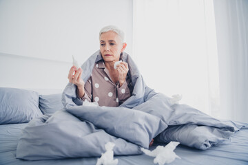 Obraz premium Charming elderly woman with white hair sitting in bed holding tissues, wearing pajamas, reflecting natural daylight