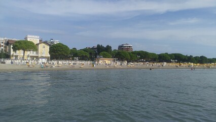 Under the pine trees on the sandy beach there are plastic deck chairs and umbrellas. People rest on them and swim in the sea. Next to the beach are buildings. Sunny autumn weather and blue sky