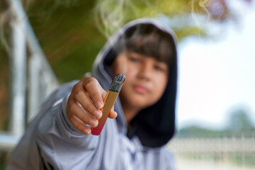 Close-up of hand holding out electronic cigarette, blurred background of a teenage boy wearing a gray hoodie over his head.