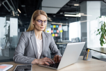 Mature woman businesswoman using laptop in modern office, focused on work. Business attire reflects...