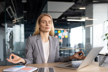 Mature woman businesswoman engaging in mindfulness meditation during office break. She sits calmly in front of laptop, seeking inner peace amidst workplace stress and daily challenges.