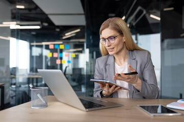 Mature woman businesswoman in office having virtual meeting using laptop and notebook for notes. Professional interaction, online collaboration, productivity in modern workspace.