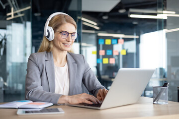 Mature woman businesswoman working online wearing headphones, focused on laptop in modern office. Engaged in virtual meeting or remote work setup, portraying professional environment and technology.