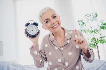 Charming elderly woman in pajamas with alarm clock in bright bedroom, smiling and cheerful