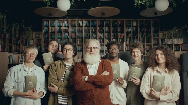 Elderly author and diverse group of young fans holding copies of book standing together in cozy modern library, looking at camera and smiling during photo session at signing event