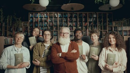 Senior author and group of young multi-ethnic readers holding copies of new book posing together for camera during meet-up in cozy modern library. Zoom shot, group portrait