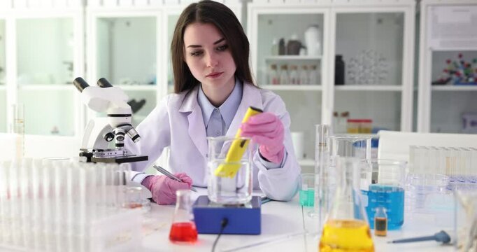 Experienced woman lab technician measures pH level of water in beaker with tool. Professional lab assistant records exact results in notebook
