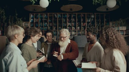 Group of young literature fans gathering around elderly author with white beard as he signing books in cozy library or bookstore during public event