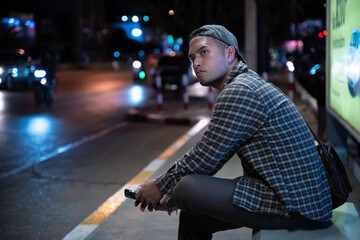 An Asian Man Waiting for a Bus After Working Late, Using His Smartphone at a Busy City Bus Stop,...