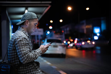 An Asian Man Waiting for a Bus After Working Late, Using His Smartphone at a Busy City Bus Stop, Capturing the Struggles of Urban Life, Overwork, and the Nightly Commute in a Chaotic Capital