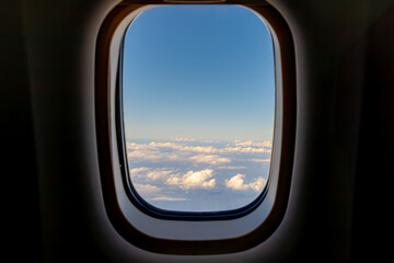 Fototapeta premium Looking through the airplane window frame with beautiful blue sky and white fluffy cloud, Aircraft flying above cumulus clouds with warm golden sunlight in the evening, Abstract nature background.