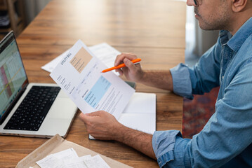 Man examining utility bill with pen and laptop at home office desk. Concept of household expenses,...