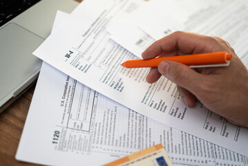 Close-up of a person reviewing tax forms with an orange pen next to a laptop. Tax preparation and financial documentation concept 