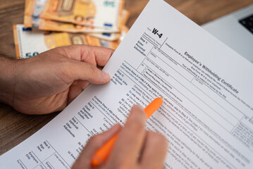 Obraz premium Close-up of a man's hands filling out a W-4 tax form with an orange pen, with euro banknotes in the background. Tax filing and financial management concept during tax season 