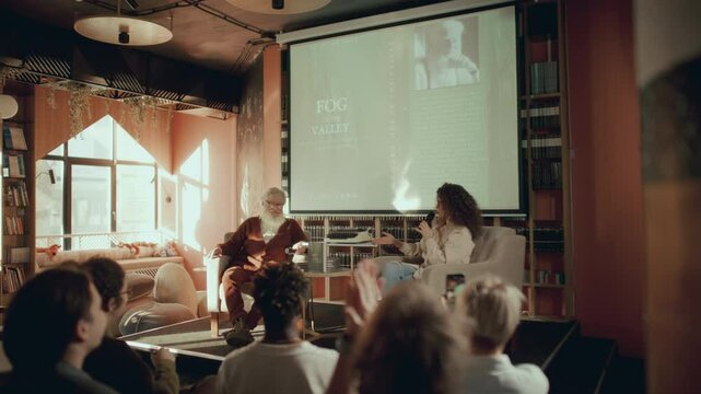 Elderly author and female moderator sitting on stage near projection screen, presenting book during literary event in modern library, as attentive audience listening and clapping hands