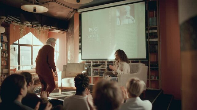 Elderly author entering sunlit library, walking on stage, greeting audience applauding him, then taking microphone and answering questions from female presenter during book launch event