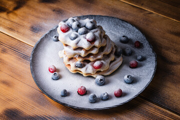 Belgian waffles with cranberries and blueberries sprinkled with powdered sugar on a wooden table on a black plate.