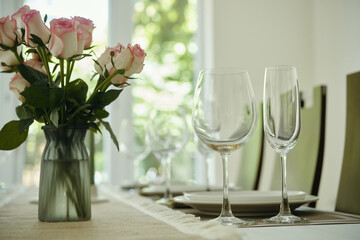 Elegant table setting featuring a vase of pink roses alongside empty wine glasses with natural light filtering through windows in background creating a serene ambiance
