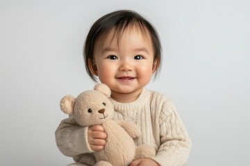 An Asian baby holding a small stuffed animal, sitting on a clean white backdrop with a gentle smile