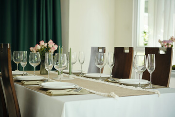 Table set for dining with glassware, white plates, and napkins arranged on a white tablecloth. Pink flowers are present in background complementing the setting