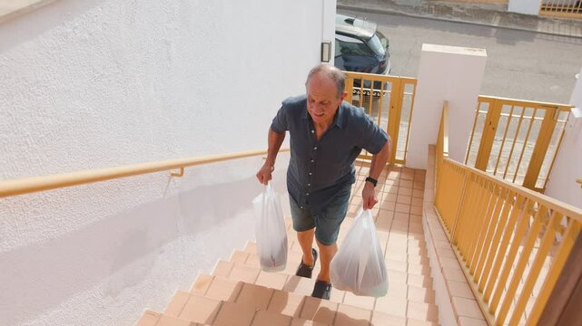 Elderly caucasian man suffers pain when carries heavy groceries bags on the exteriors stairs of the house. Stressed and ache expression. Frontal view, following shot, hight angle, horizontal 4k.