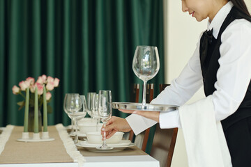 Person arranging dinnerware and glassware on a table adorned with flowers, preparing for a formal gathering. Careful attention is given to detail, ensuring perfect placement