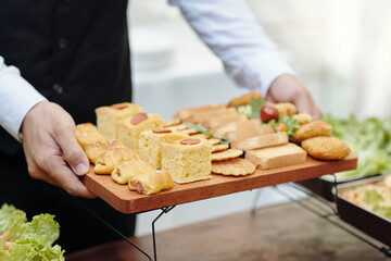 Close-up view of hands holding tray with various appetizers, including sandwiches and pastries. Tray is served during outdoor event, with leafy greens in the background