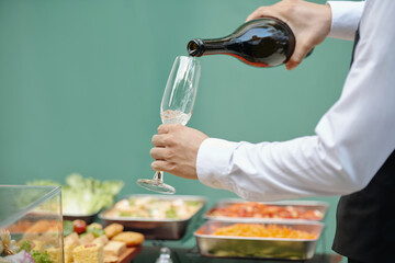 Close-up of person holding wine bottle and pouring wine into glass in an outdoor catering event with various dishes on display