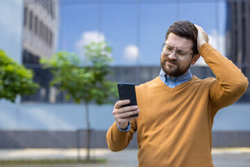 Upset and disappointed young man standing on the street and looking at the mobile phone, holding...