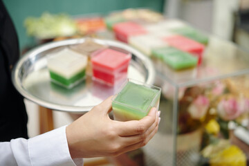 Hand holding tray with colorful cubes, possibly soap or jelly, in a market or shop. Bright colors and background with floral arrangements adding vibrant feel