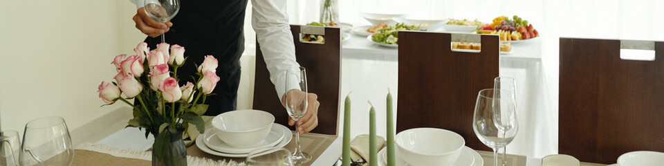 Person in white shirt and vest arranging glassware on table adorned with pink roses, plates, and napkins while a variety of food is visible in background