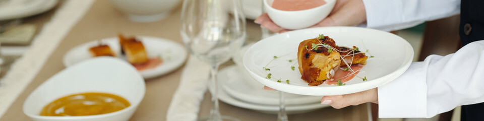 Closeup of server holding beautifully plated gourmet dish garnished with microgreens. Table set with elegant white dishes, glassware, and decorative napkins creating a refined dining experience