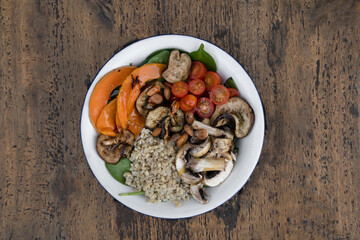 Top view of a nutritious salad with grilled vegetables. Vegetarian salad with grilled pumpkin, mushrooms, spinach, barley, almonds and cherry tomatoes, in a white bowl on the restaurant wooden table.	