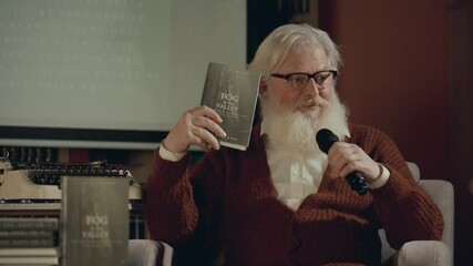 Senior writer with long white beard sitting on stage in front of audience, holding his book and talking into microphone during book launch or lecture