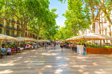 Tourists walk past sidewalk cafes on the famous La Rambla street through the historic Mediterranean city of Barcelona, Catalonia, Spain. 