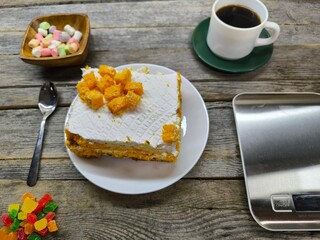 There is a delicious piece of cake on the plate next to a cup of coffee and marshmallows. Calorie counting scales
