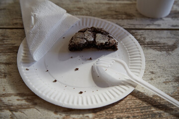 A paper plate with one half of a chocolate biscuit with cracks and a fork in a beam of sunlight on a wooden table, close-up, unfinished breakfast, diet, limiting sweet and pastry consumption.