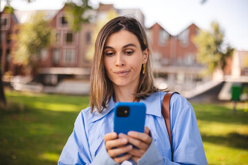 Woman holding a phone. Expressive puzzled woman thinking outside. Woman in doubt with smartphone in her palm, think about reading information, search solution. Girl wear shirt, bag, top, walk in park.